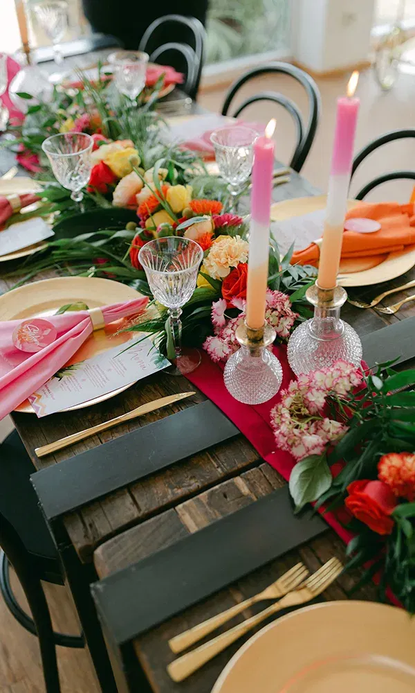 Elegant gedeckter Tisch mit goldenen Tellern, Kristallgläsern und pinkfarbenen Kerzen, die in dekorativen Vasen leuchten. Die Blumenarrangements in kräftigen Farben setzen Akzente auf dem rustikalen Holztisch.