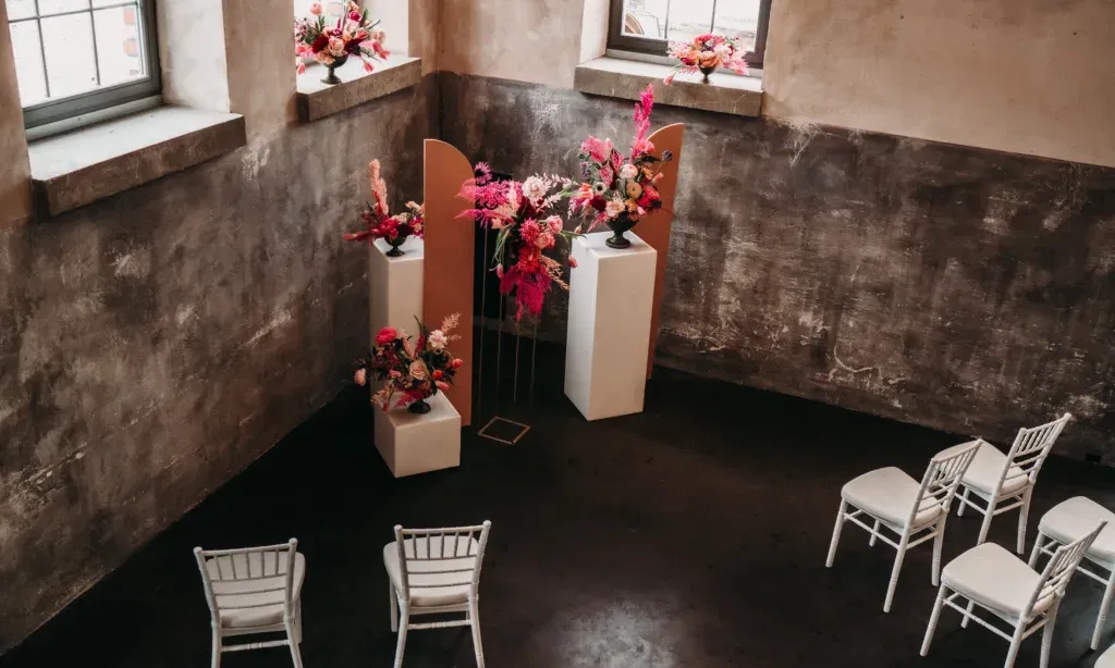 Eine minimalistische Hochzeitsdekorationsszene in einem rustikalen Innenraum mit rauen, grauen Wänden. In der Ecke stehen weiße Sockel mit farbenfrohen Blumengestecken in kräftigen Rosa- und Orangetönen. Auf den Fensterbänken befinden sich ebenfalls florale Arrangements, die den Raum lebendiger machen. Weiße, klassische Stühle stehen in Reihen, die eine intime Atmosphäre für die Zeremonie schaffen. Der gesamte Stil wirkt modern und elegant, mit einem Hauch von Industrial-Chic.