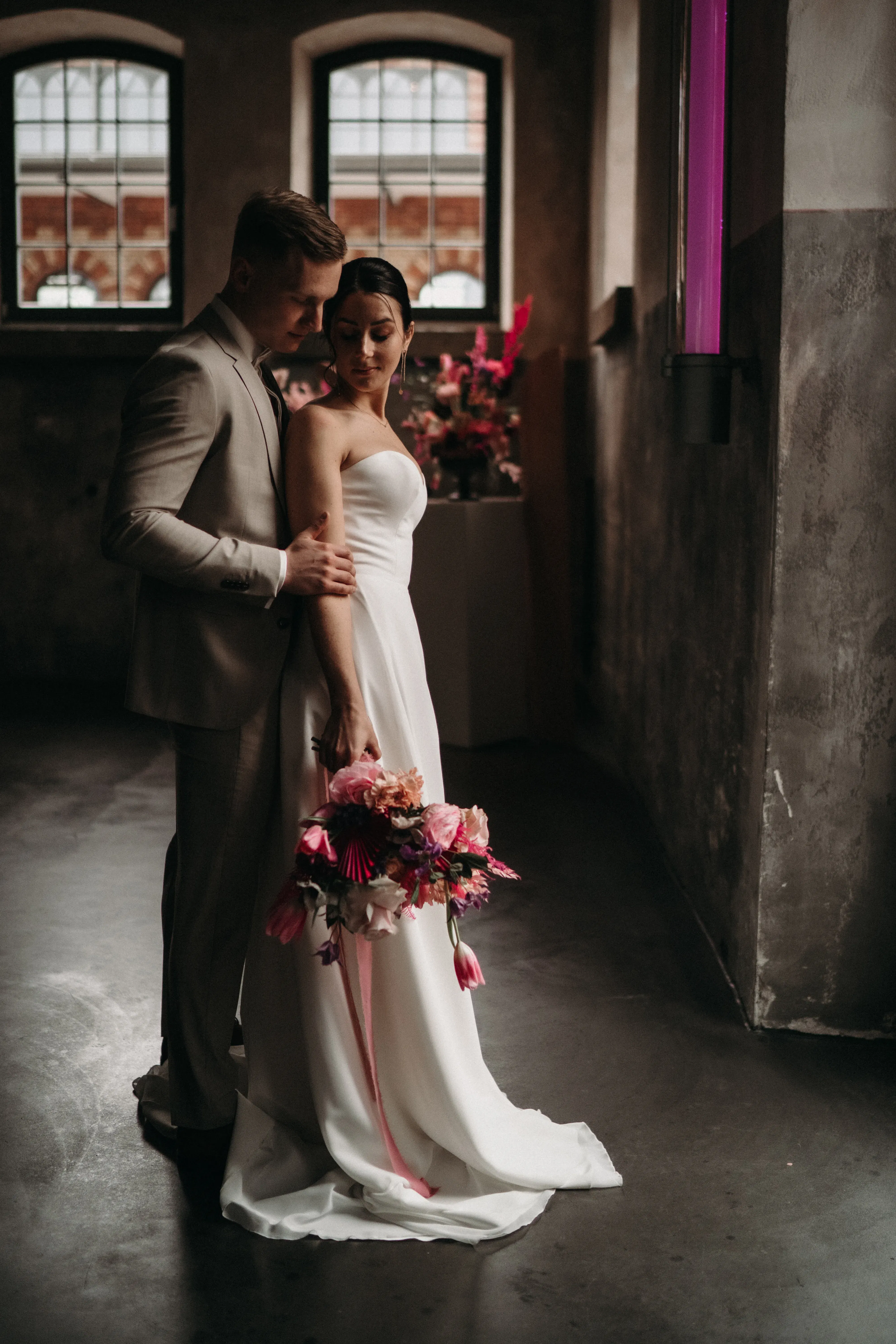 Das frisch verheiratete Paar posiert in einem minimalistischen Raum vor zwei großen Fenstern. Die Braut trägt ein elegantes weißes Kleid und hält einen opulenten Blumenstrauß, der Bräutigam steht dicht hinter ihr in einem grauen Anzug und berührt zärtlich ihre Schulter. Der Raum ist in gedämpftem Licht, wodurch der Blumenstrauß in kräftigen Farben hervorsticht.