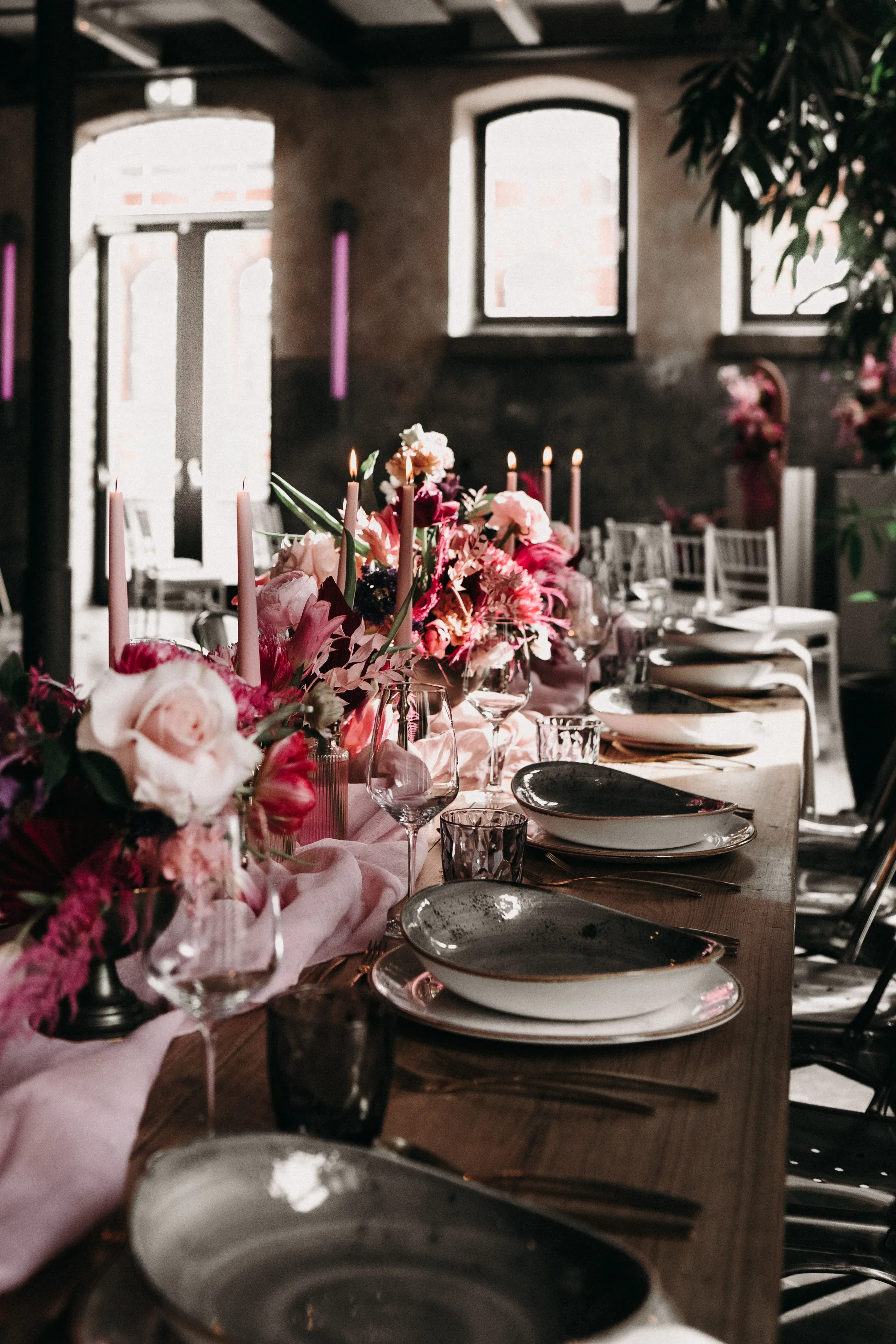 Eine lange Tafel ist für ein festliches Dinner gedeckt, mit rustikalen Tellern, feinem Glasgeschirr und einer opulenten Blumendekoration in kräftigen Pink- und Rottönen. Mehrere Kerzen brennen entlang der Tischmitte, umrahmt von einer zarten, rosa Stoffbahn, während Tageslicht den Raum durch hohe Fenster durchflutet.