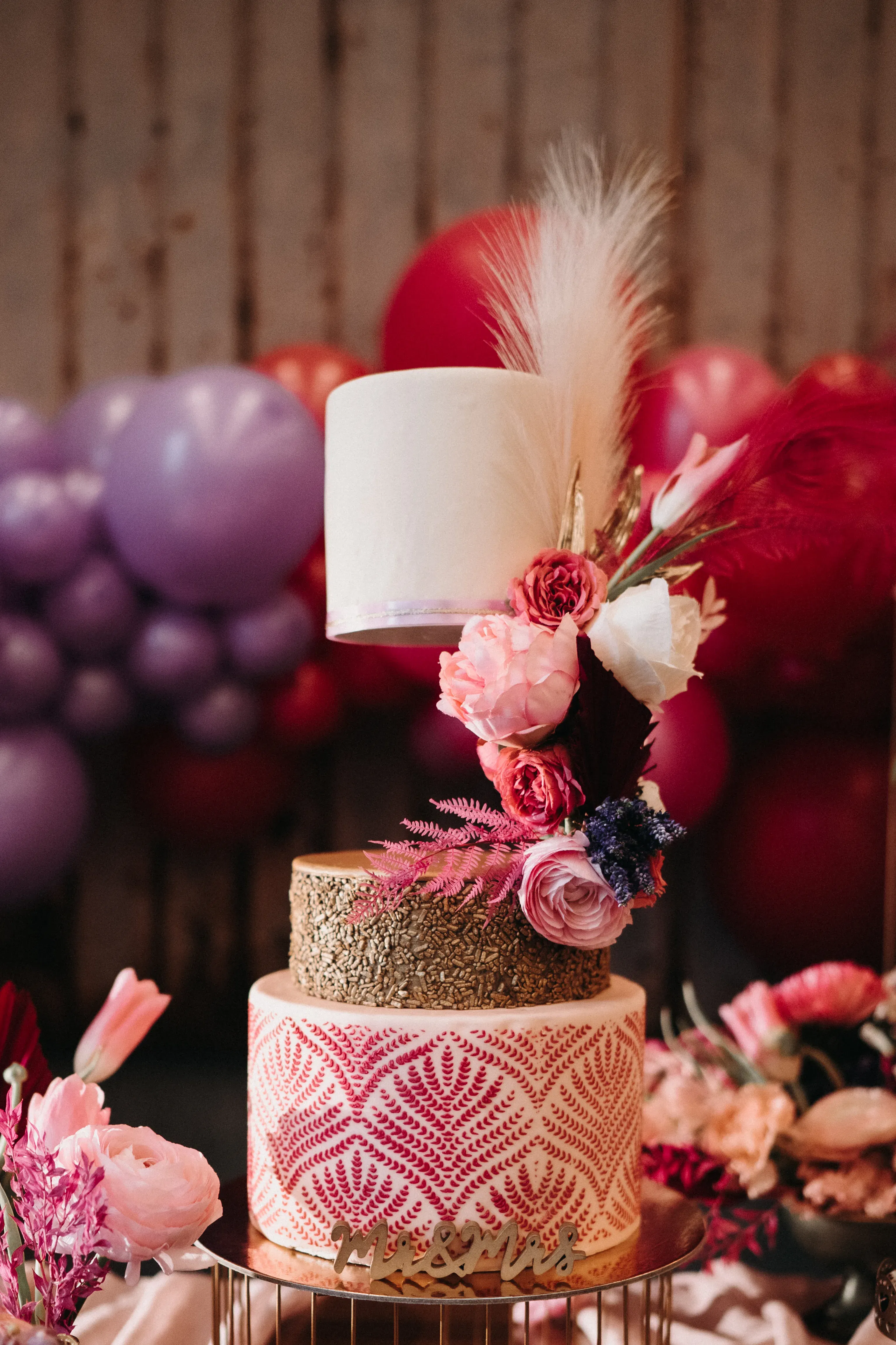 Detailaufnahme einer kunstvoll gestalteten Hochzeitstorte mit drei Schichten. Die untere Schicht ist mit einem filigranen rosa Muster versehen, die mittlere goldfarben und die obere in schlichtem Weiß gehalten. Bunte Blumen in Rosa und Pink zieren die Torte, die vor einer festlichen Dekoration aus bunten Luftballons präsentiert wird.