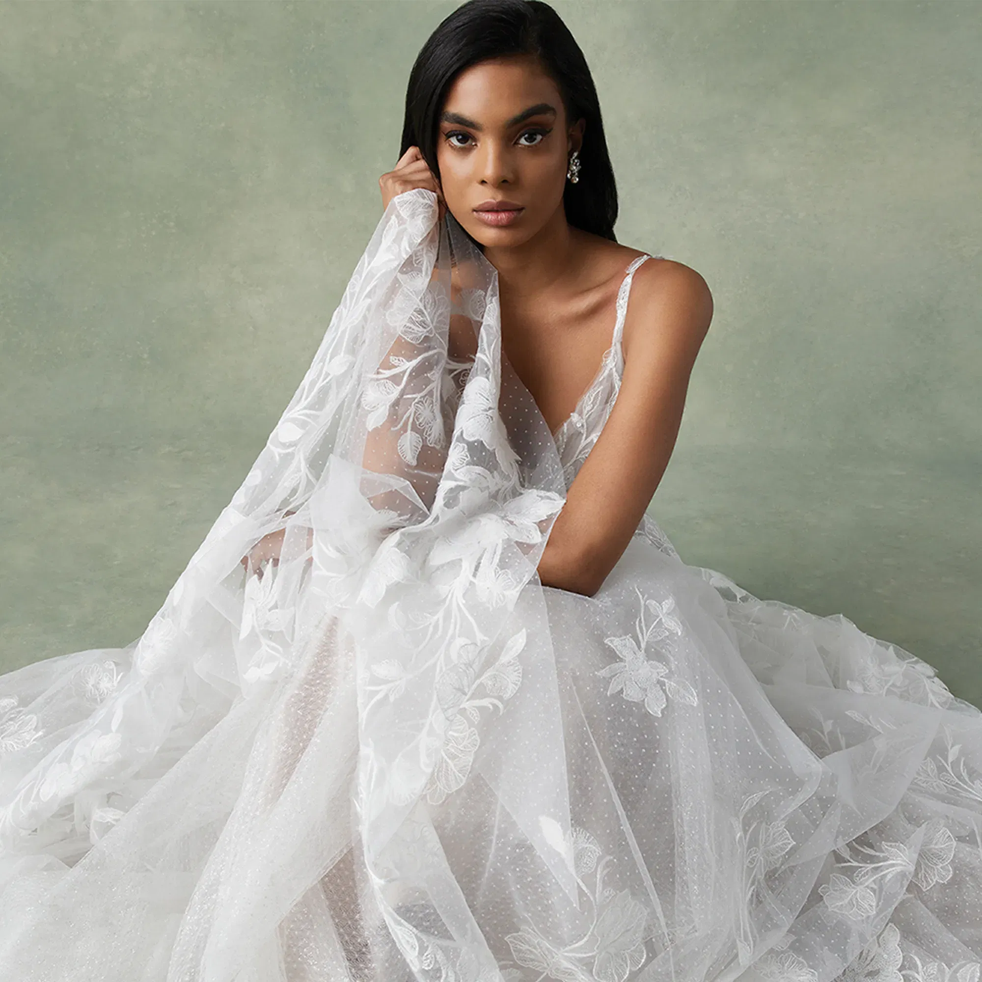 Eine stilvolle Braut mit dunklem, glattem Haar trägt ein fließendes Brautkleid aus Tüll und Spitze, während sie anmutig ihren Schleier hält. Der weiche, grüne Hintergrund hebt die zarte Textur des Kleides hervor.