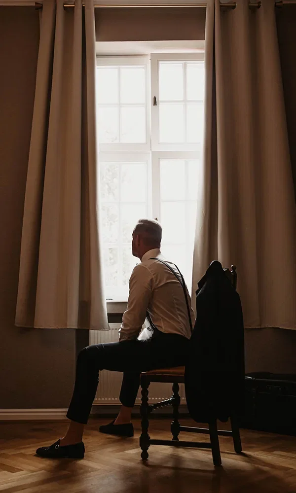 Ein Bräutigam sitzt vor einem Fenster und schaut hinaus. Bekleidet mit einem weißen Hemd und Hosenträgern, ist seine Silhouette vom Tageslicht eingerahmt. Sein schwarzes Jackett liegt über der Stuhllehne.