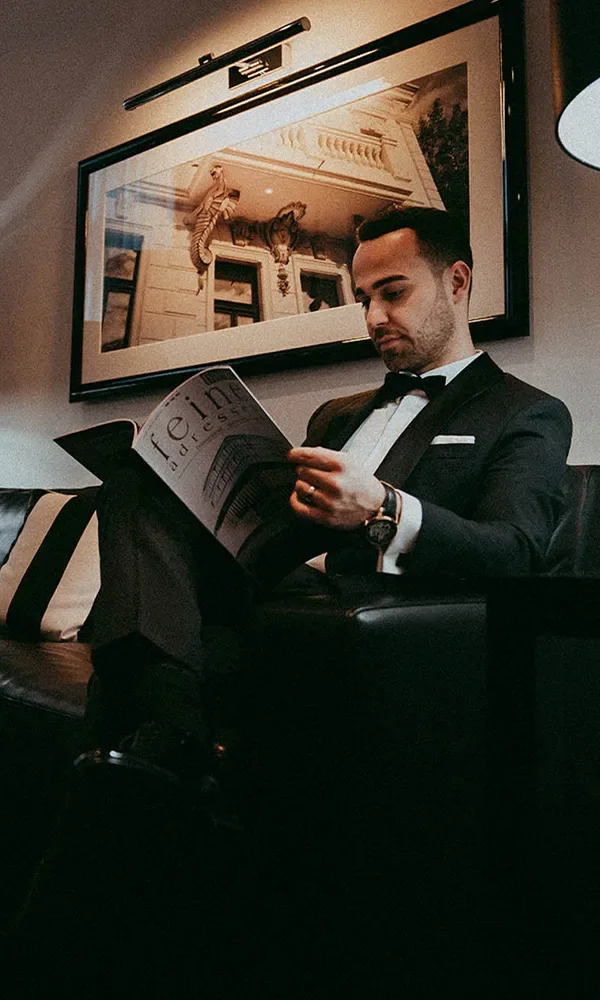 Ein Mann im Smoking sitzt entspannt auf einem schwarzen Ledersofa und blättert durch ein Magazin. Über ihm hängt ein großes Kunstwerk an der Wand, und die warme Beleuchtung verleiht dem Raum ein luxuriöses Ambiente.
