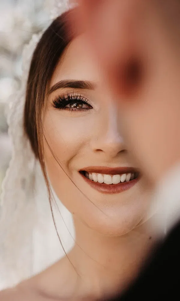 Eine Braut lächelt in einer Nahaufnahme, während ein verschwommener Teil eines Mannes im Vordergrund zu sehen ist. Ihr Make-up hebt ihre Augen mit langen Wimpern und natürlichem Lippenstift hervor, sie trägt einen Schleier.