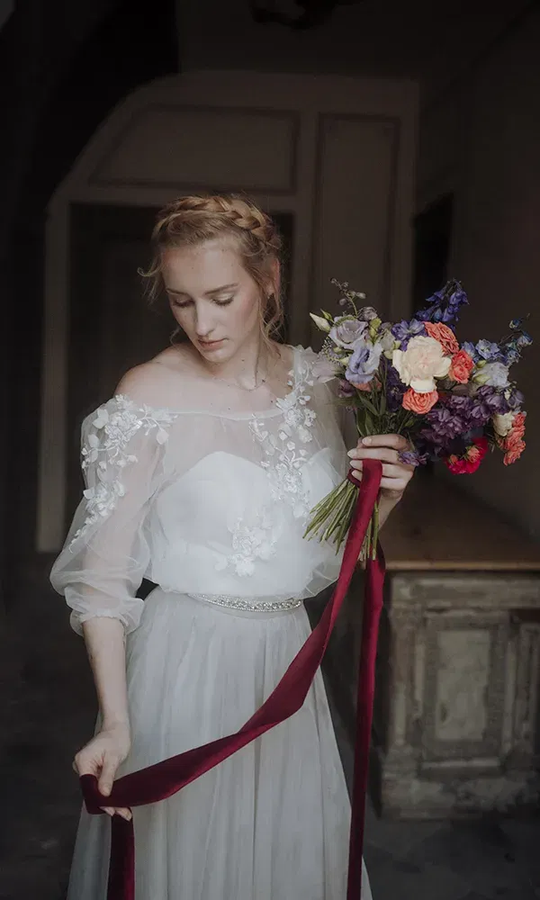 Eine Braut in einem zarten, floralen Hochzeitskleid hält einen Blumenstrauß mit roten und violetten Blumen. Ein rotes Band hängt von ihrem Strauß, während sie nachdenklich auf den Boden schaut.