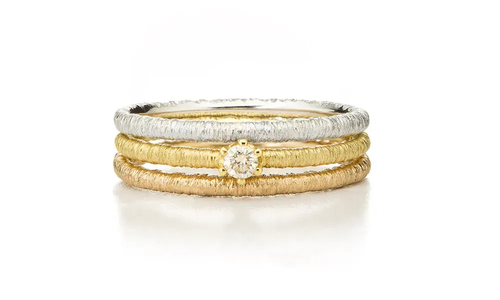 Drei schmale Ringe in unterschiedlichen Edelmetallen liegen übereinander. Der mittlere Ring ist mit einem funkelnden Diamanten geschmückt, während die Oberfläche der Ringe eine feine Struktur aufweist, die an handgewebte Textilien erinnert.