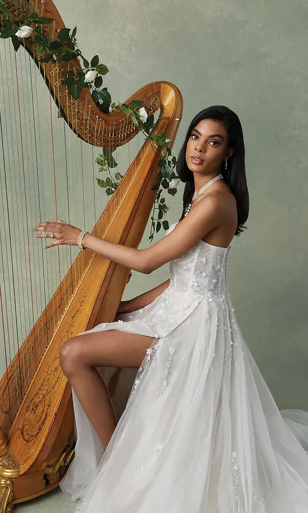 Die Braut posiert neben einer mit Blumen geschmückten Harfe und trägt ein detailreiches, weißes Kleid mit Perlenstickerei. Sie sitzt aufrecht, und ihr Blick ist ruhig und anmutig. Die Szene wird durch die floralen Verzierungen an der Harfe romantisch abgerundet.