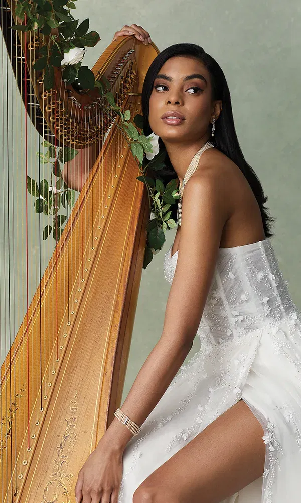 Eine Frau in einem glamourösen weißen Brautkleid sitzt anmutig vor einer kunstvoll verzierten Harfe, die von Blumen und grünen Blättern geschmückt ist. Sie trägt dezenten Schmuck und blickt mit einem ausdrucksvollen, nachdenklichen Gesichtsausdruck in die Ferne. Das Bild strahlt Eleganz und Ruhe aus.