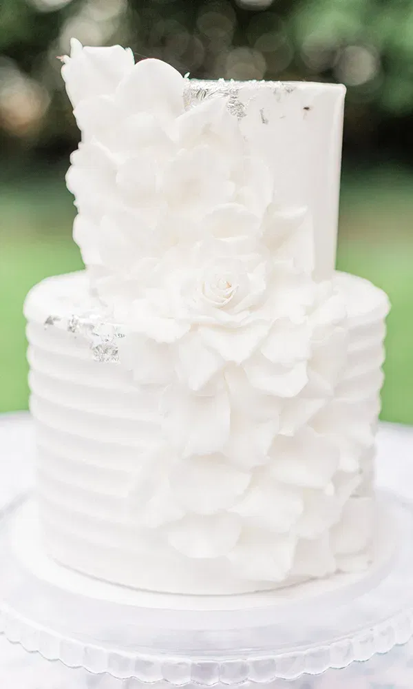 Die weiße Hochzeitstorte ist mit eleganten Blüten und silbernen Akzenten verziert. Sie steht auf einem durchsichtigen Tortenständer und präsentiert sich in einem Garten mit grünem Hintergrund.
