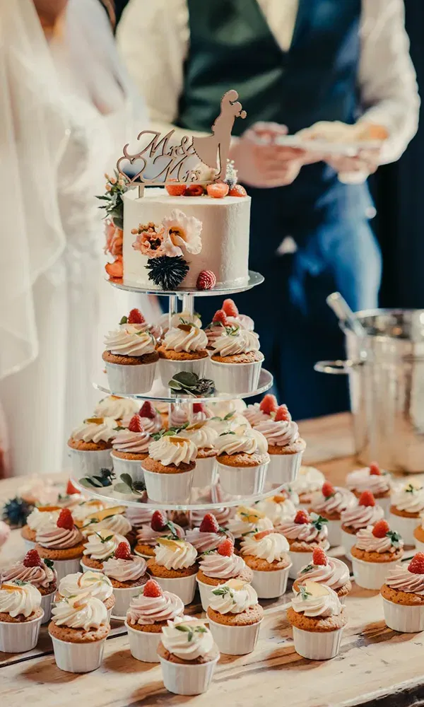 Eine Hochzeitstorte steht auf einem Cupcake-Turm, dekoriert mit frischen Erdbeeren und Sahne. Im Hintergrund sind unscharf das Brautpaar zu erkennen, während die Torte den Mittelpunkt des Tisches bildet.