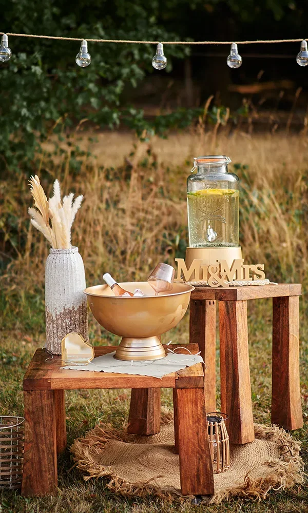 Ein rustikal dekorierter Getränketisch im Freien. Eine goldene Schale mit Flaschen auf Eis steht auf einem Holztisch, während ein Glasgefäß mit Limonade daneben steht. Der Tisch ist mit einer "Mr & Mrs"-Dekoration und Trockenblumen verziert.