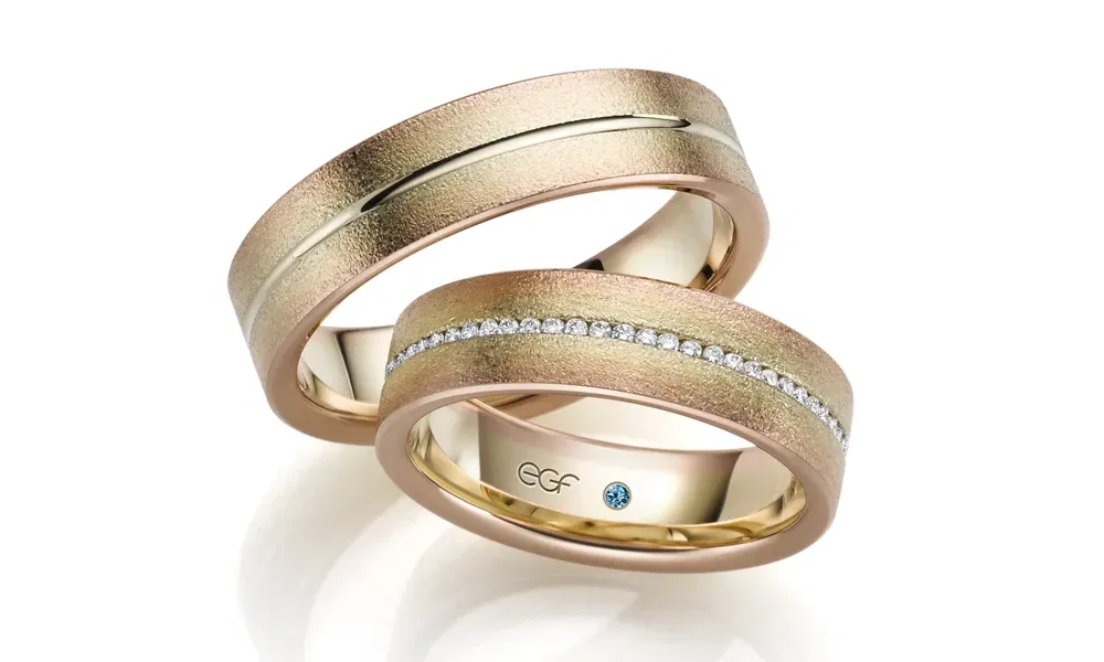 Zwei elegante, breite Eheringe aus Rosé- und Gelbgold mit einem gebürsteten Finish. Der Damenring ist in der Mitte mit glitzernden Diamanten verziert, während der Herrenring eine dezente Linie auf der Oberfläche trägt. Die Ringe sind schlicht und doch luxuriös gestaltet.