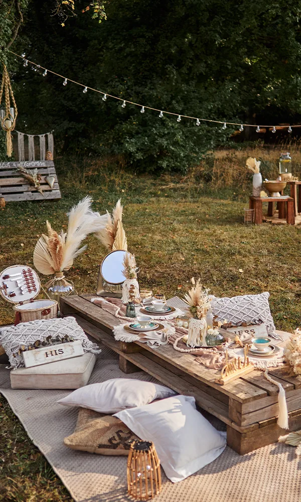 Ein rustikal gedeckter Holztisch im Freien, umgeben von weichen Kissen und Decken. Auf dem Tisch befinden sich Blumen, Geschirr und Kerzen, während eine Schaukel und eine mit Lichterketten dekorierte Szenerie den Hintergrund füllen.