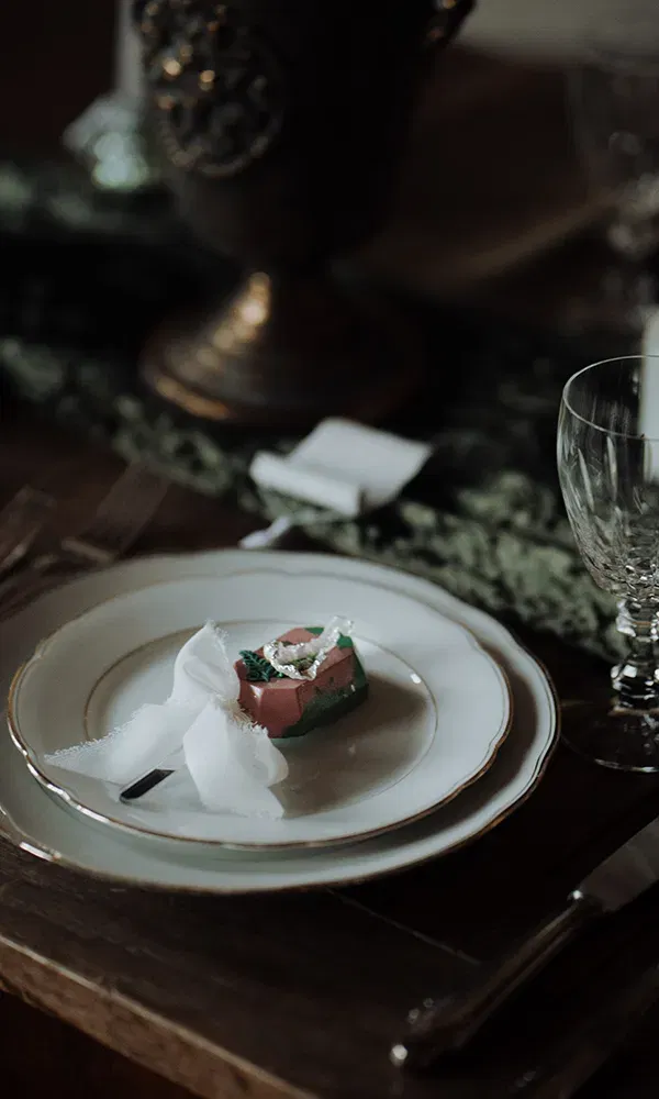 Auf einem weißen Teller liegt ein farbiges Gastgeschenk, das mit einem durchsichtigen Stoffband umwickelt ist. Die elegante Tischdekoration umfasst Besteck, ein Weinglas und Kerzen.