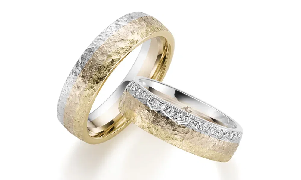 Zwei massige Eheringe mit einer auffälligen Kombination aus Gelb- und Weißgold. Der Ring der Braut glänzt durch eine Reihe von Diamanten, die entlang des oberen Randes eingesetzt sind, während beide Ringe durch eine gehämmerte, rustikale Oberfläche bestechen.