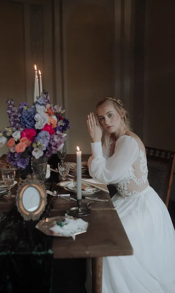 Die Braut sitzt an einem gedeckten Tisch mit silbernen Kerzenhaltern und einem üppigen Blumenarrangement aus violetten und roten Blumen. Sie schaut ruhig in die Kamera, während die Kerzen brennen.