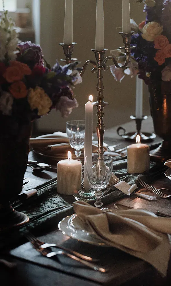 Ein elegant dekorierter Tisch, auf dem Kerzen in verschiedenen Größen und Blumenarrangements zwischen Silberkerzenständern stehen. Gläser und Geschirr sind sorgfältig um die Dekoration herum angeordnet.