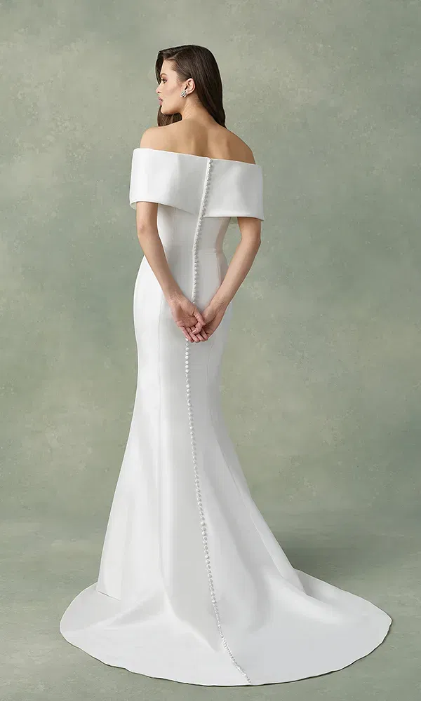 Die Rückansicht eines schlichten und dennoch raffinierten Brautkleides, dessen schulterfreies Design die obere Rückenpartie der Braut zur Geltung bringt. Eine durchgehende Knopfleiste zieht sich von den Schultern bis zum Saum des fließenden Kleides, das auf dem Boden aufliegt.