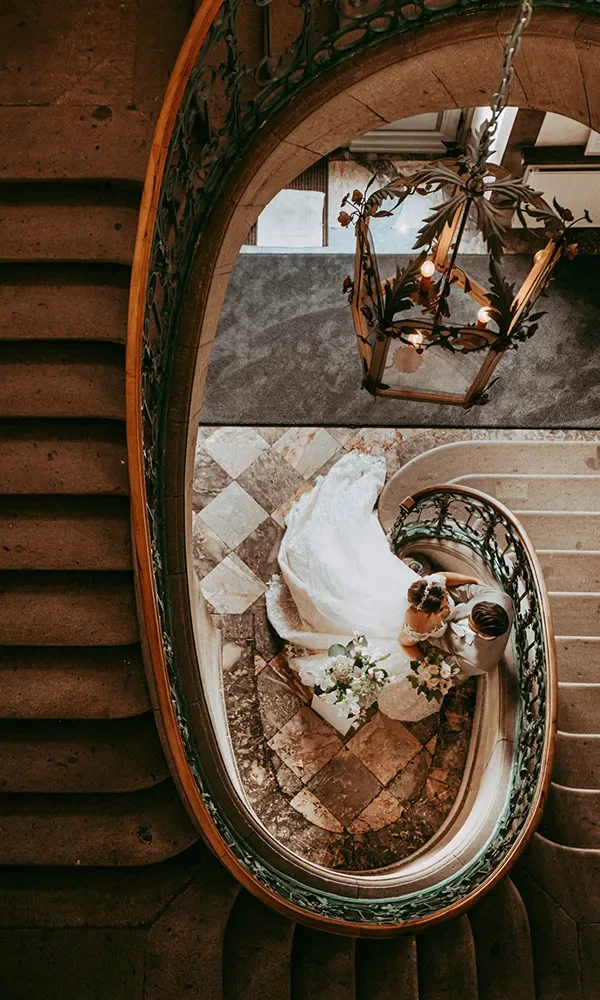 Blick von oben auf eine Braut und einen Bräutigam, die auf einer gewundenen Treppe stehen. Der Boden unter ihnen ist mit Marmorfliesen bedeckt, und eine große Hängelampe ist über ihnen sichtbar.