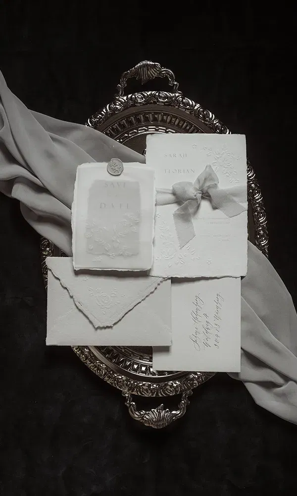Hochzeitseinladungen und Briefumschläge mit geprägten Blumenmustern sind auf einem reich verzierten Silbertablett platziert, das von einem grauen Tuch umrahmt wird.