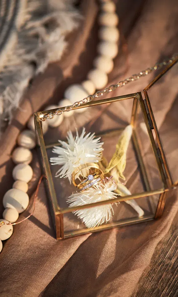Goldene Eheringe mit einem Diamanten sind in einer Glasbox mit einer zarten Trockenblume platziert. Die Box steht auf einem braunen Tuch, und im Hintergrund ist eine Holzkette als Dekoration sichtbar.