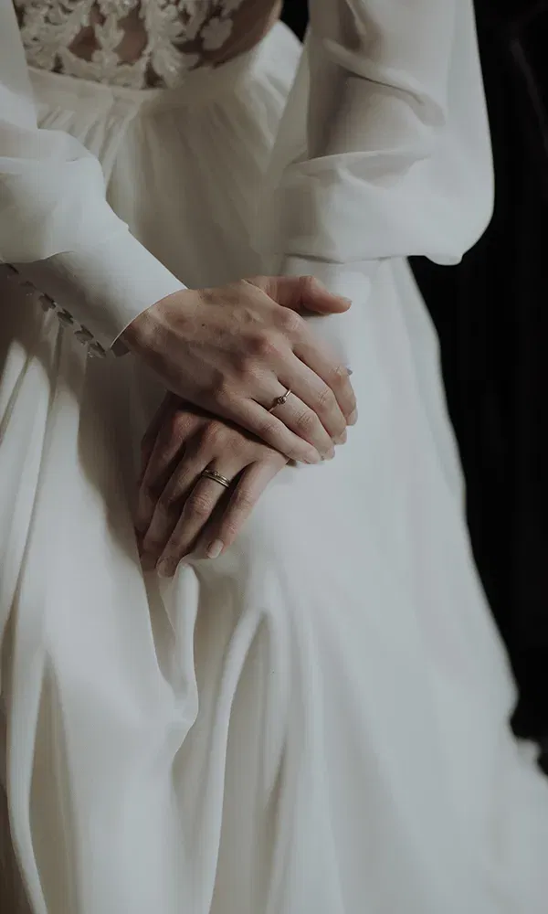 Die gefalteten Hände der Braut ruhen sanft auf ihrem Hochzeitskleid. Ihr Ringfinger trägt einen schlichten Ehering, und das elegante Kleid mit Spitze ist im Detail zu erkennen.