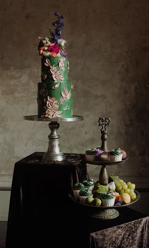Eine aufwendig verzierte Hochzeitstorte in Grün, geschmückt mit Blüten, steht auf einem Tisch. Neben ihr befindet sich eine Etagere, die mit Cupcakes und Früchten wie Trauben und Limetten gefüllt ist.