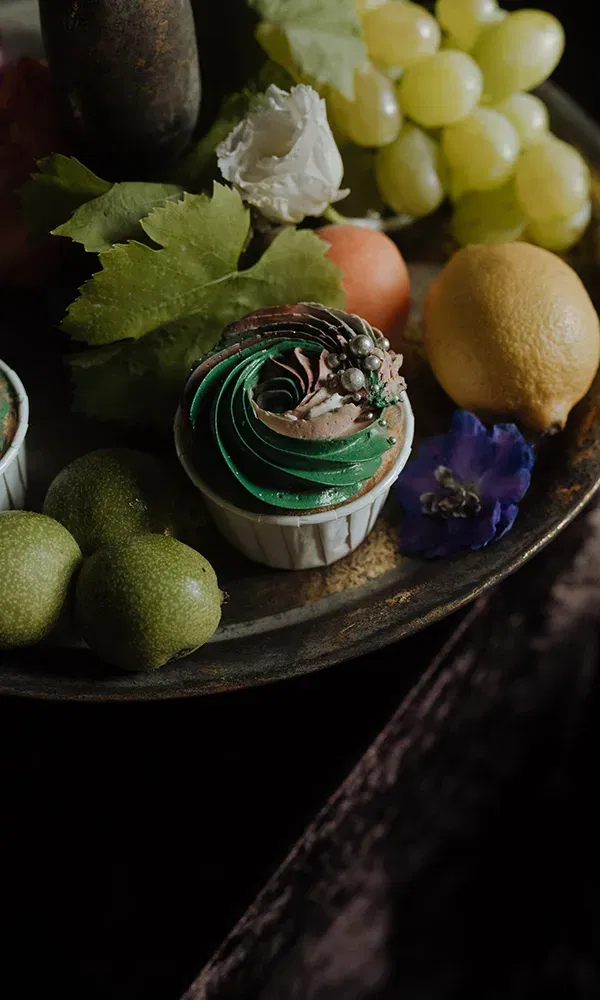 Auf einem Tablett liegt ein dekorierter Cupcake mit grünem Zuckerguss, umgeben von verschiedenen Früchten wie Trauben, Limetten und einer Zitrone. Blumen und Blätter runden die Komposition ab.