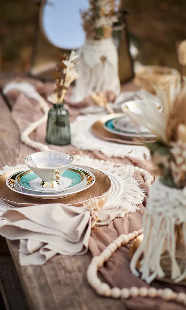 Ein rustikaler Holztisch ist mit elegantem Geschirr und einer goldenen Unterlage gedeckt. Trockenblumen in einer grünen Vase sowie geflochtene Tischdekorationen verleihen dem Arrangement einen Boho-Stil.