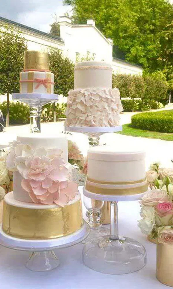 Mehrere Hochzeitstorten mit goldenen Akzenten und floralen Verzierungen sind auf einem Tisch arrangiert. Jede Torte hat ein individuelles Design und steht auf einem gläsernen Tortenständer.