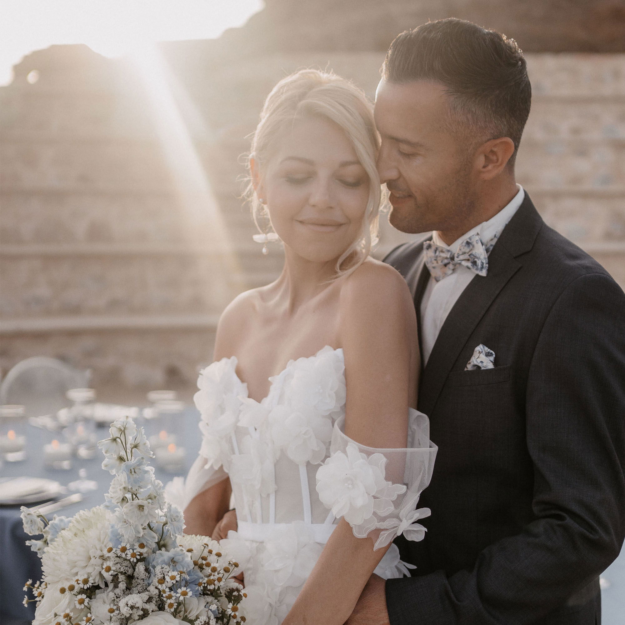 Eine romantische Szene mit einem Brautpaar, das sich eng umarmt. Die Braut trägt ein elegantes weißes Kleid mit Blumenapplikationen und einen zarten Blumenstrauß, während der Bräutigam einen dunklen Anzug und eine gemusterte Fliege trägt. Die Abendsonne wirft weiche Strahlen auf das Paar, das den Moment in Stille genießt.
