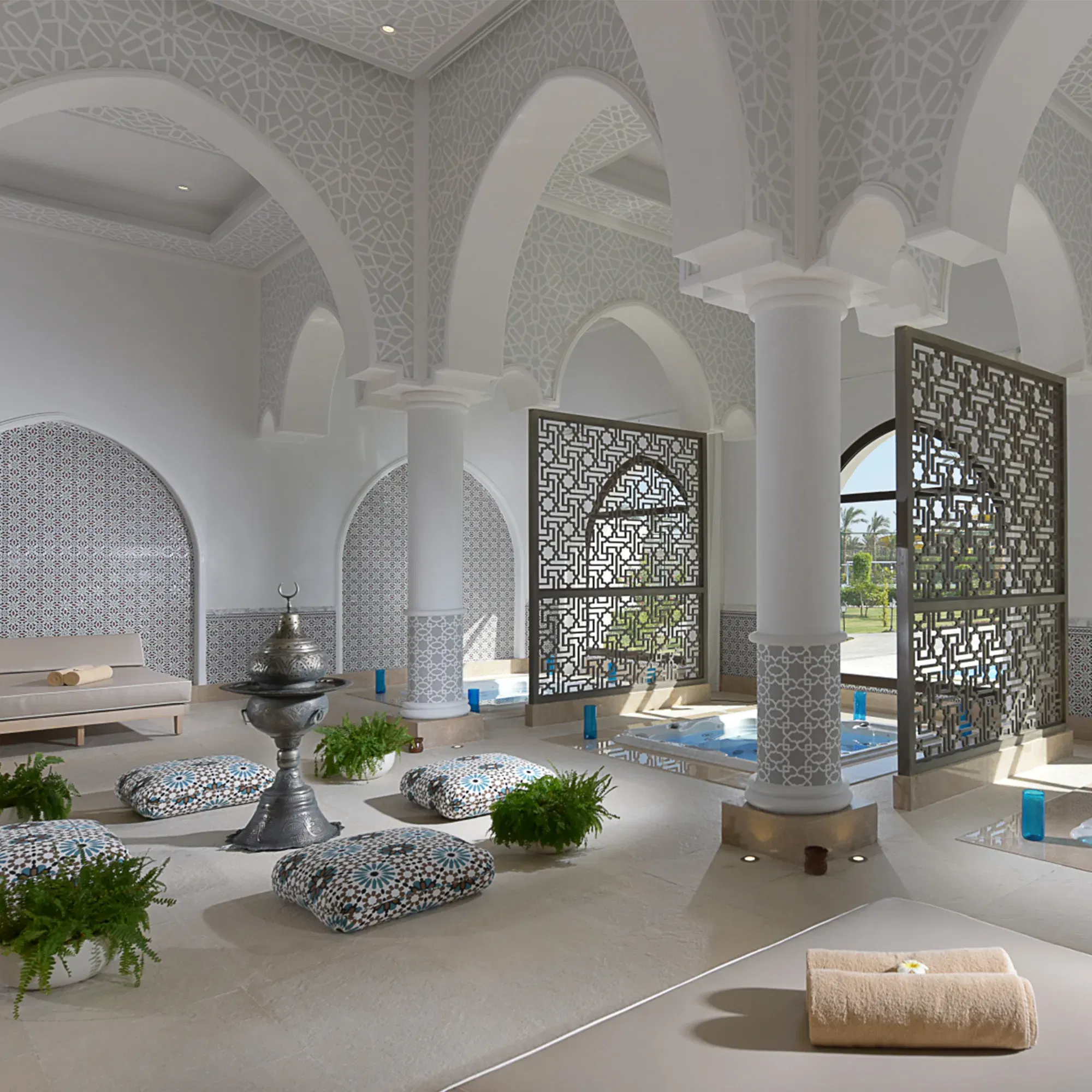 Ein elegantes Spa mit orientalischer Architektur, ausgestattet mit Säulen, kunstvollen Trennwänden und gemütlichen Sitzkissen. Der Raum ist hell und friedlich, mit einem zentralen Brunnen und dezenter Dekoration.