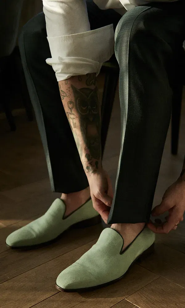 Der Bräutigam, dessen Arm mit einem Eulentattoo verziert ist, zieht sich elegante, hellgrüne Slipper an. Die Szene spielt sich auf einem Holzboden ab, und die Ärmel seines weißen Hemdes sind hochgekrempelt.