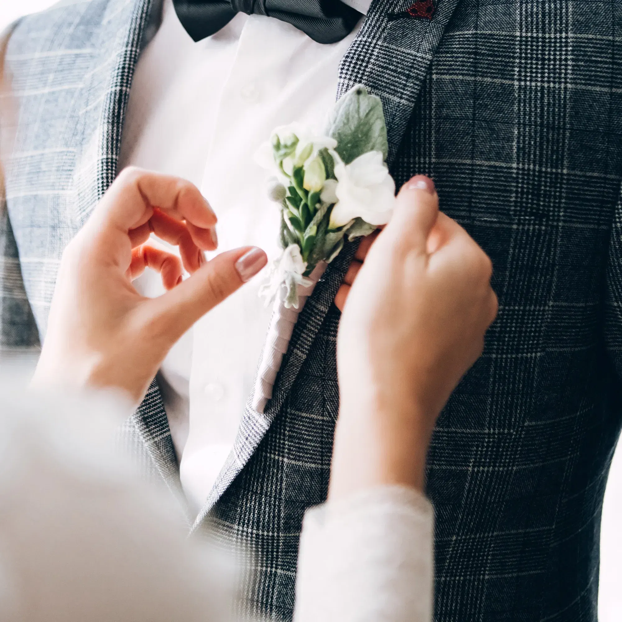 Eine Hand richtet die Boutonnière des Bräutigams, der einen eleganten, karierten Anzug mit weißem Hemd und schwarzer Fliege trägt. Die weiße Blume rundet den festlichen Look ab.