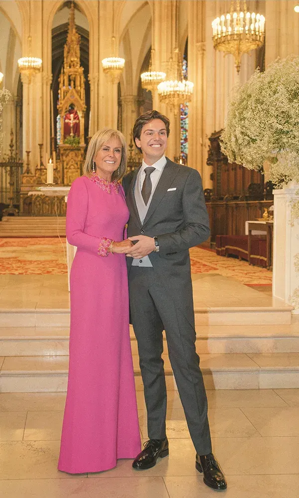 Der Bräutigam posiert stolz mit seiner Mutter, die ein auffälliges pinkfarbenes Kleid trägt, in einer historischen Kirche mit Kronleuchtern und einem kunstvollen Altar im Hintergrund.