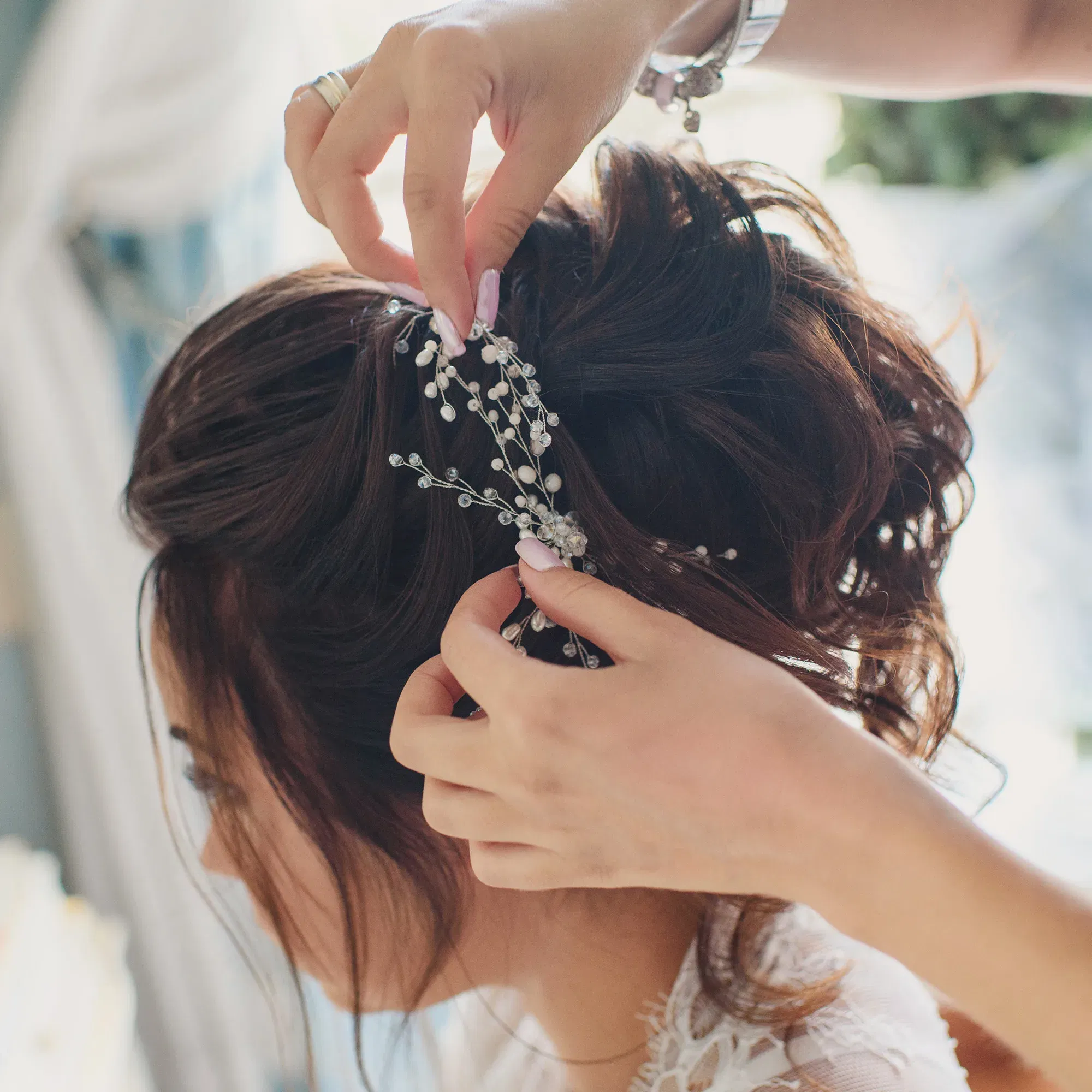 Hände stecken eine verzierte Haarspange mit Perlen und Kristallen in die elegante Hochsteckfrisur der Braut. Ihr Haar ist kunstvoll hochgesteckt und passt perfekt zur festlichen Stimmung.