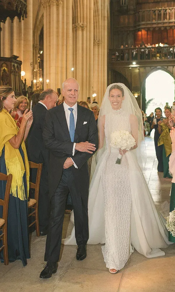 Der Vater führt seine Tochter, die Braut, den Gang einer prunkvollen Kirche entlang. Sie trägt ein weißes Brautkleid mit Schleier, während die Gäste den Einzug mit Handys und Lächeln begleiten.