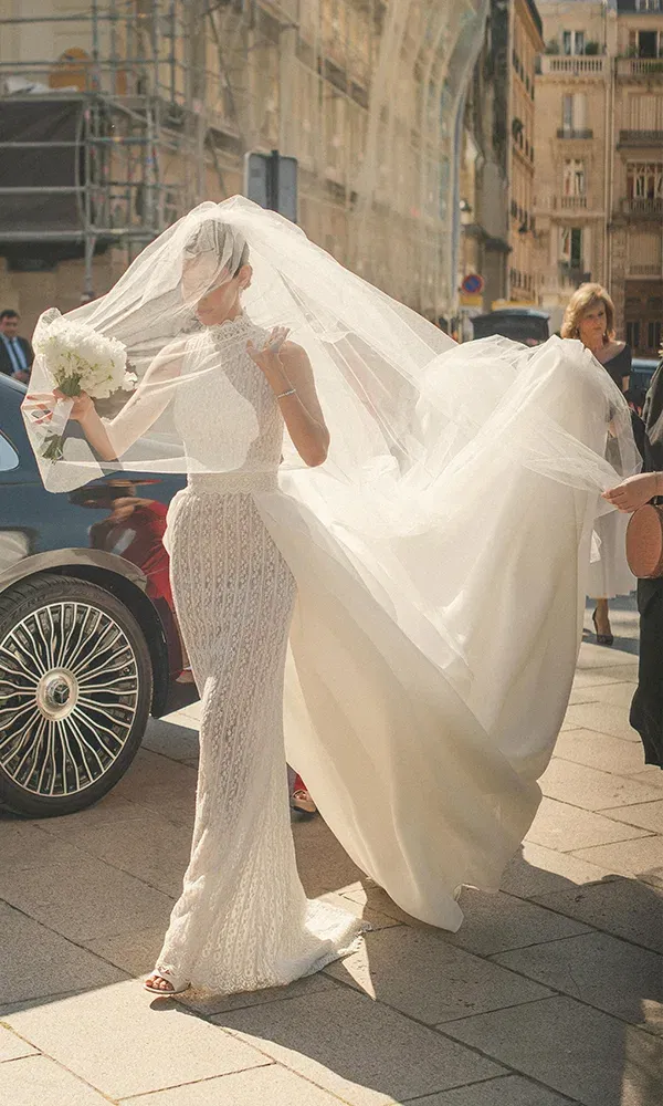 Eine stilvolle Braut in einem hochgeschlossenen, perlenbestickten Hochzeitskleid wird von ihrem langen Schleier umhüllt, während sie aus einem schwarzen Auto steigt und ihren Blumenstrauß trägt.