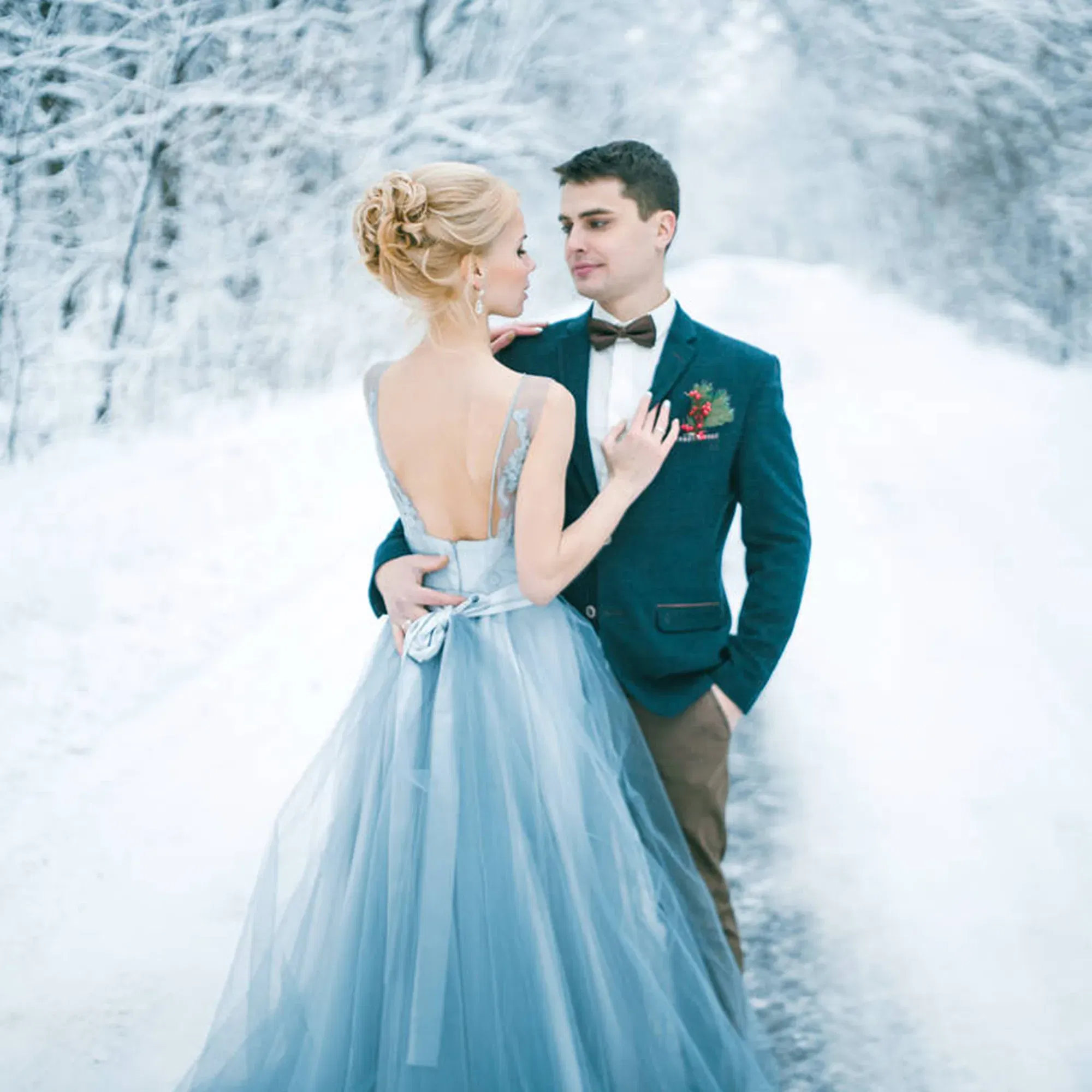 Inmitten einer winterlichen Szenerie steht ein Paar auf einem schneebedeckten Weg. Die Frau trägt ein blaues Kleid mit Schleifendetail, während der Mann sie in einem schicken Blazer und einer Fliege zärtlich umarmt.