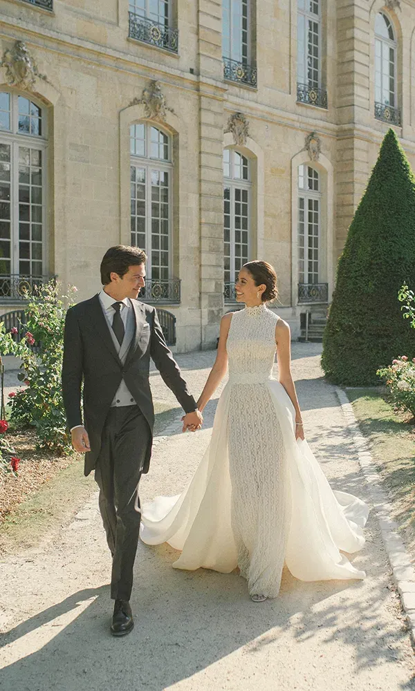 Die Braut und der Bräutigam gehen Arm in Arm durch die Gärten eines prächtigen Schlosses. Die Braut trägt ein hochgeschlossenes Brautkleid mit langem Schleier, und der Bräutigam trägt einen klassischen Hochzeitsanzug.