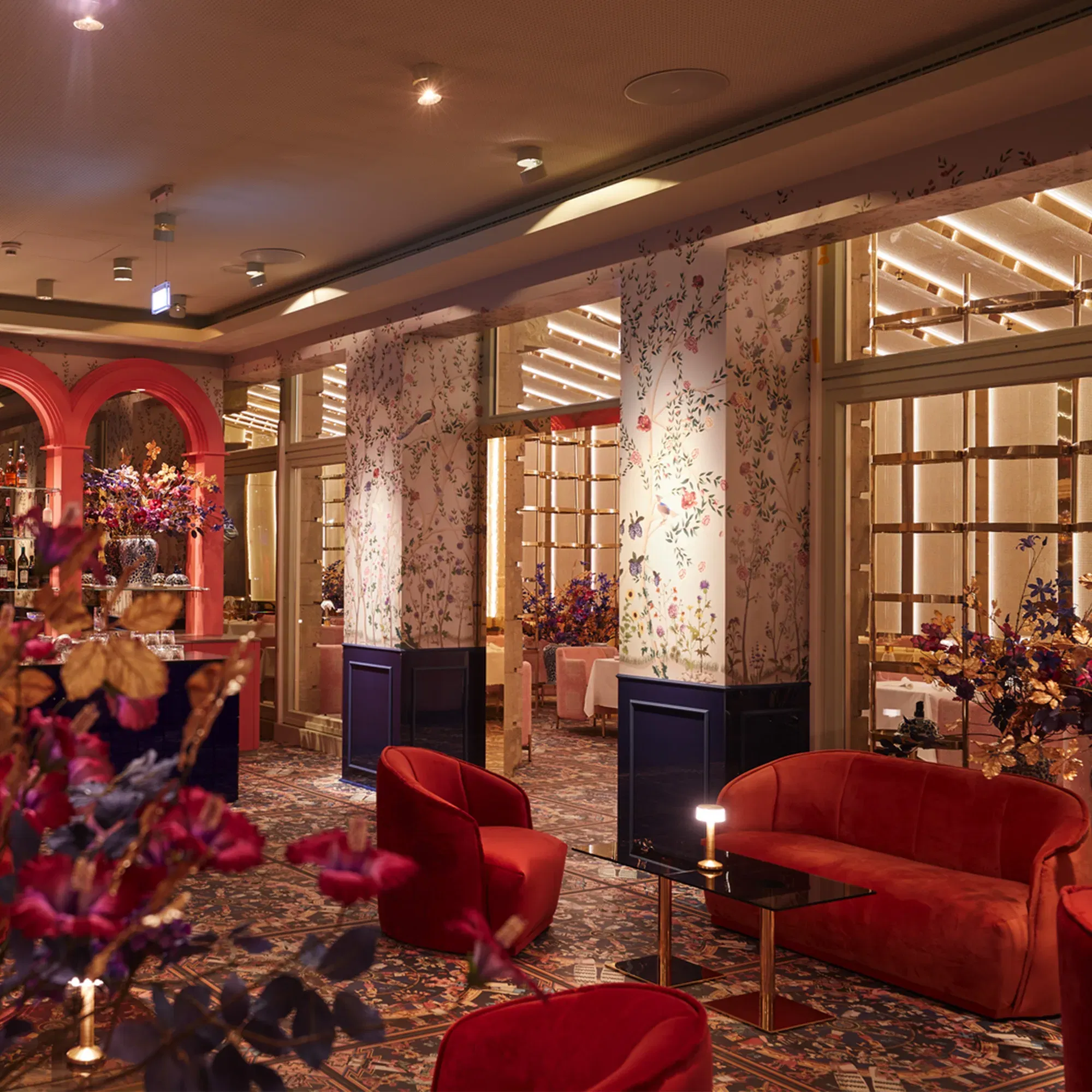 Ein stilvoll eingerichteter Raum mit roten Samtstühlen und dekorativen Blumen in intensiven Farben. Die mit Blumenmustern verzierten Säulen und die warme Beleuchtung verleihen dem Raum Eleganz und Intimität.