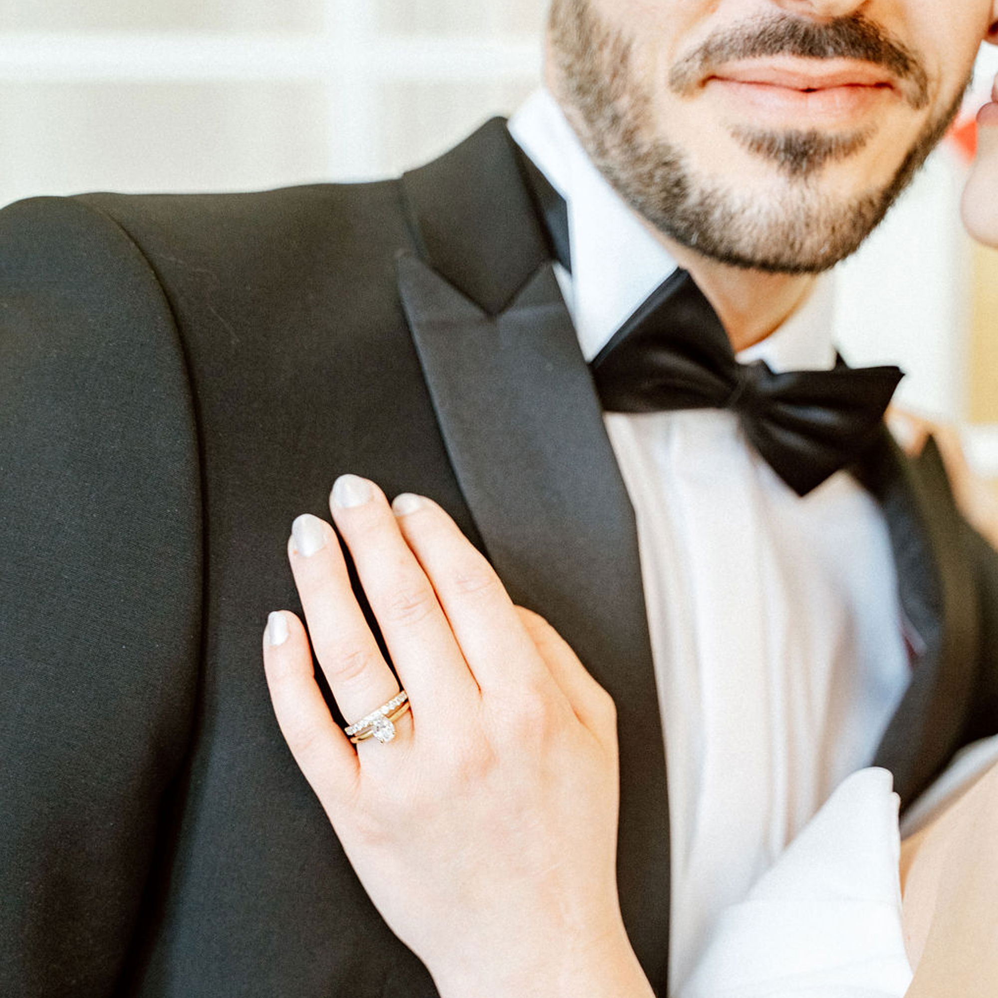 Detailansicht einer Brauthand, die den Smoking des Bräutigams berührt, wobei der funkelnde Verlobungsring auf ihrem Finger besonders hervorsticht. Der Bräutigam trägt eine klassische schwarze Fliege.
