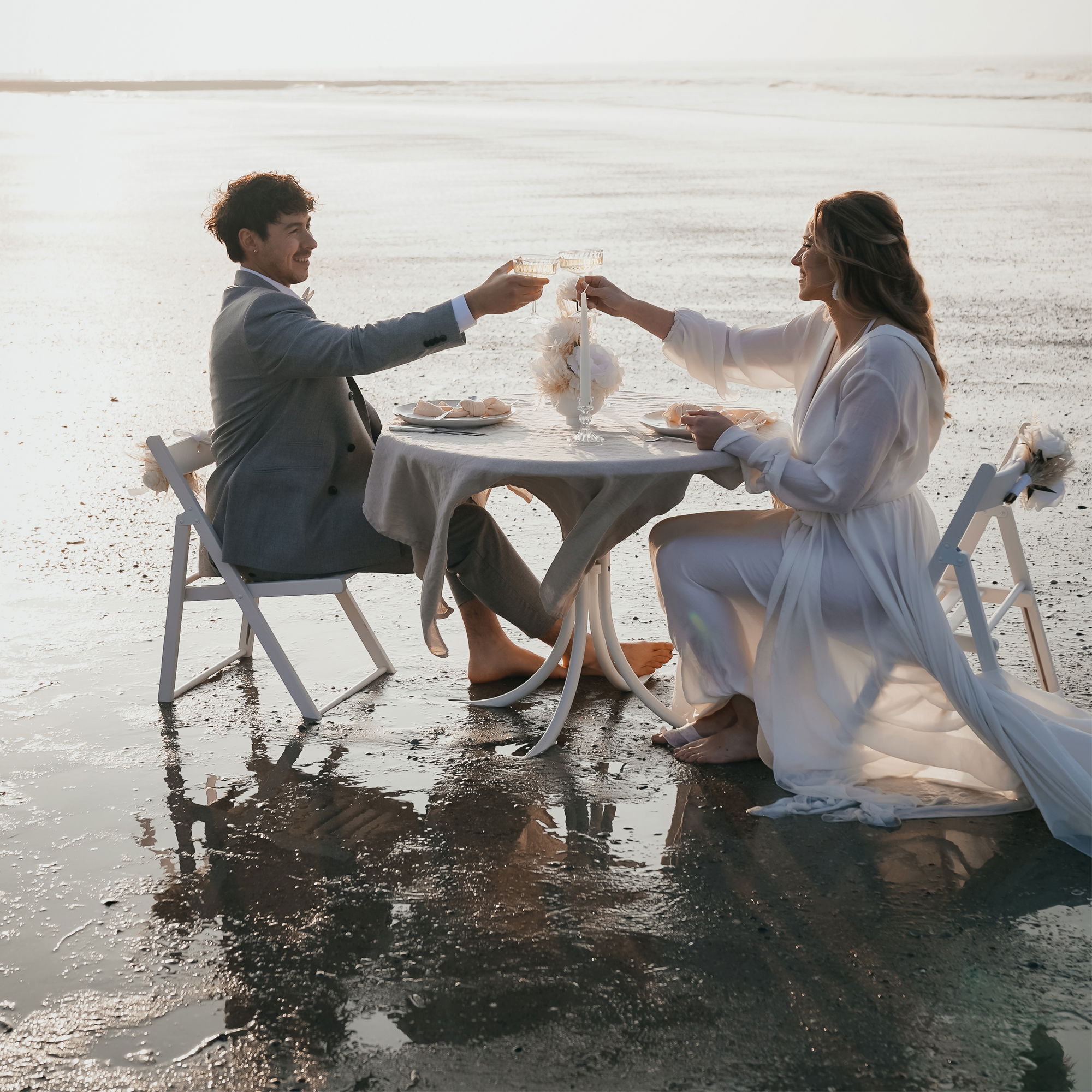 Braut und Bräutigam feiern ihre Hochzeit am Strand, während sie barfuß an einem Tisch sitzen und mit Champagnergläsern anstoßen. Die Abendsonne spiegelt sich auf dem nassen Sand.