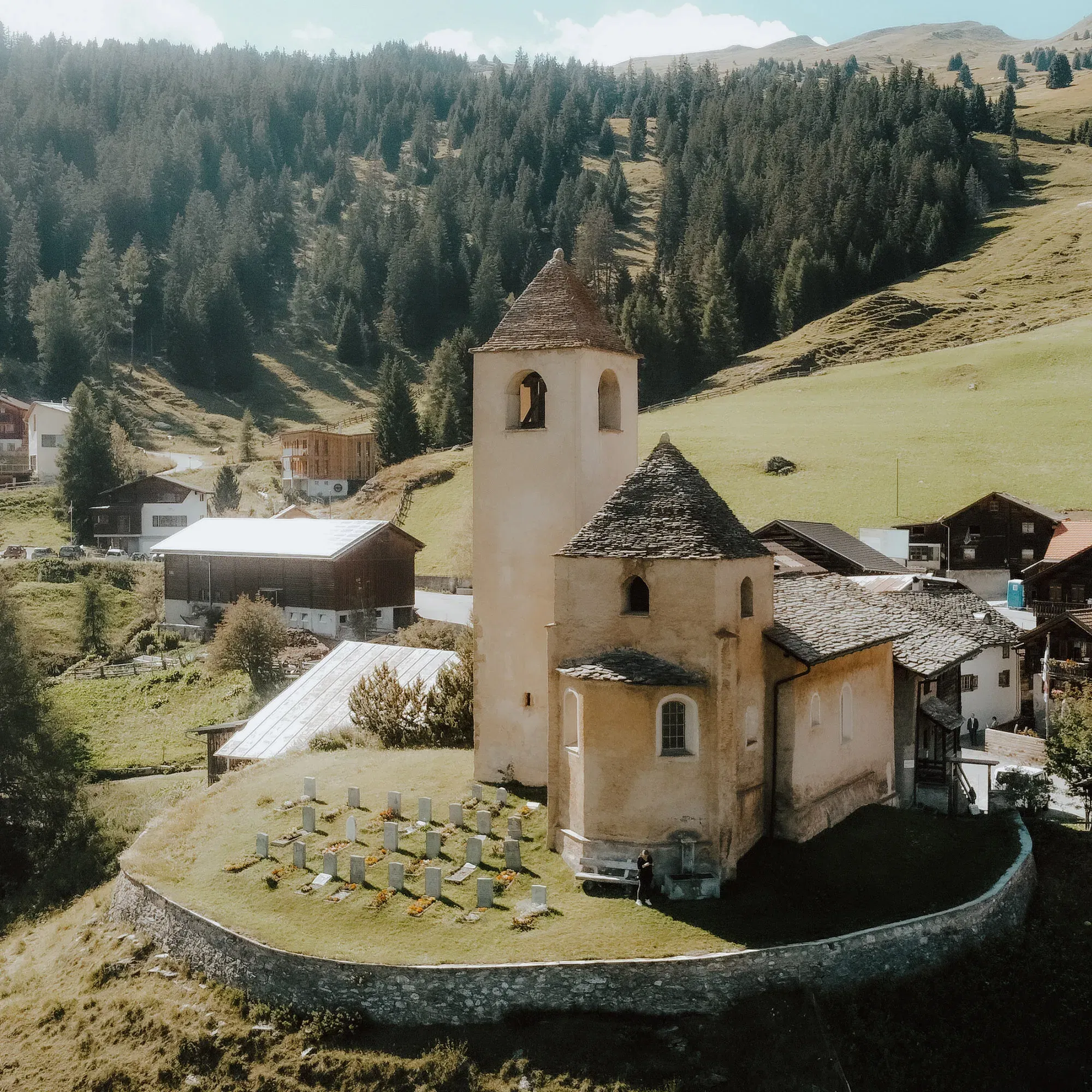 Eine historische Kirche in ländlicher Umgebung, umgeben von Bergen und Wäldern, mit einem Friedhof und traditionellen Gebäuden in der Nähe.