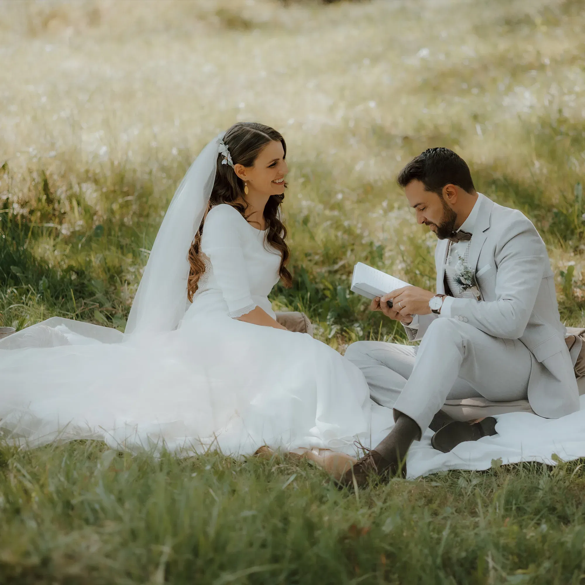 Inmitten einer grünen Wiese sitzt das Brautpaar und teilt einen intimen Moment, während der Bräutigam einen Brief vorliest. Die Braut trägt ein weißes Kleid mit Schleier, und der Bräutigam ist in einen grauen Anzug gekleidet.