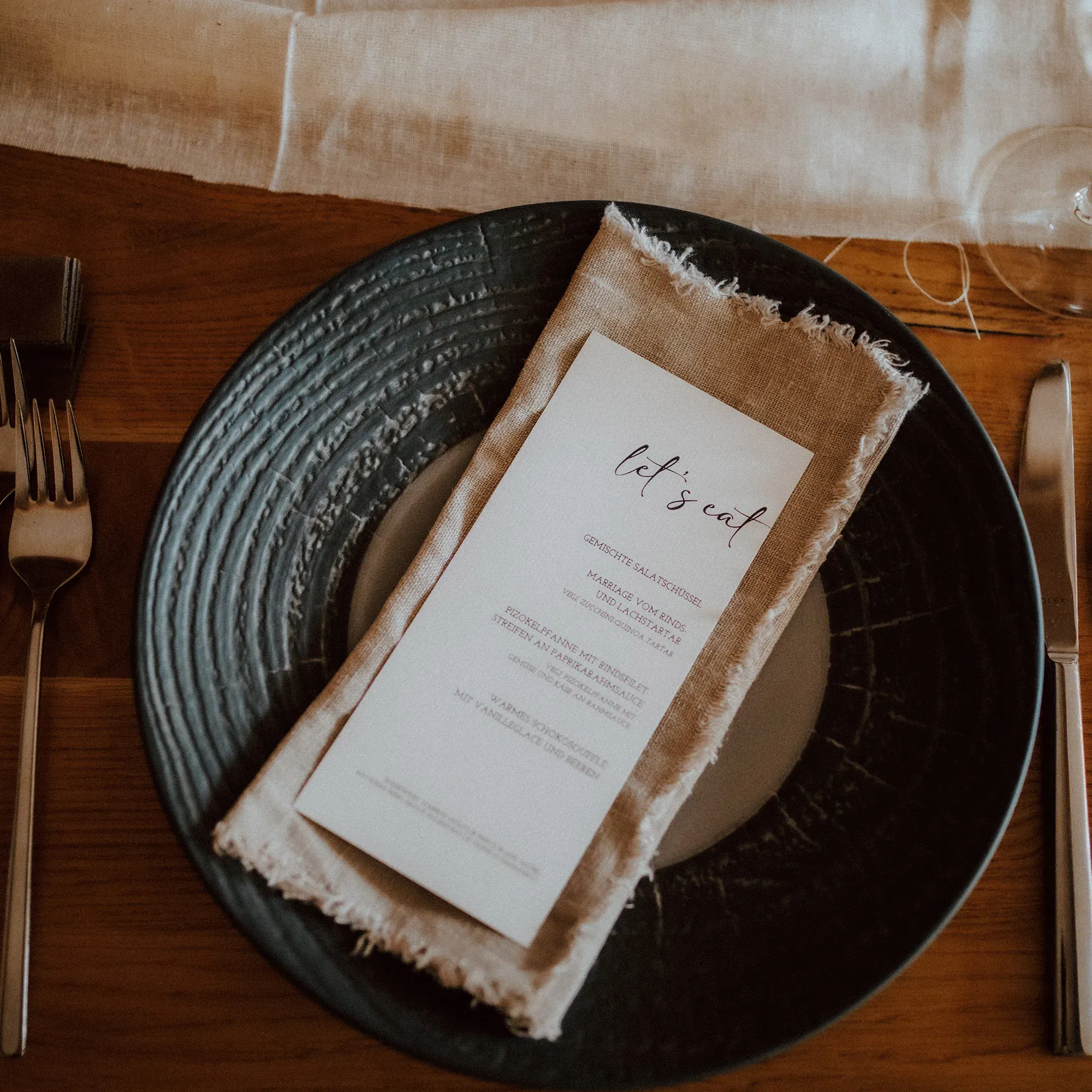 Ein moderner Hochzeitstisch, bei dem eine rustikale Serviette auf einem Keramikteller liegt, dazu eine Menükarte, die das Hochzeitsessen ankündigt.