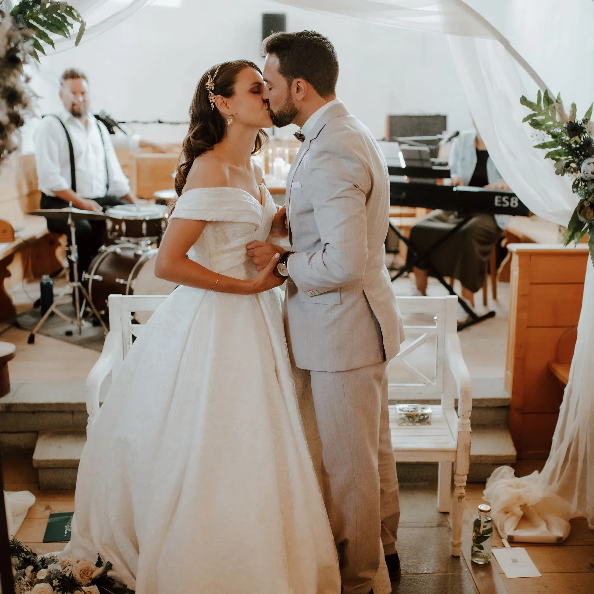 Das Brautpaar teilt einen romantischen Kuss während ihrer Trauung, die von einer Band musikalisch begleitet wird. Die Braut trägt ein elegantes Kleid, der Bräutigam einen hellen Anzug, während ein Schlagzeuger und ein Keyboarder im Hintergrund zu sehen sind.