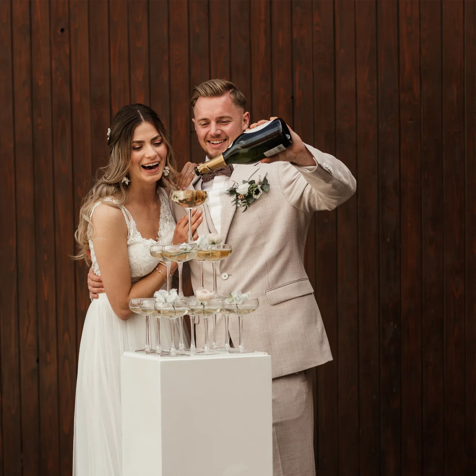 Braut und Bräutigam füllen bei ihrer Hochzeitsfeier eine Pyramide aus Champagnergläsern. Die Braut strahlt, während der Bräutigam die Flasche hält.