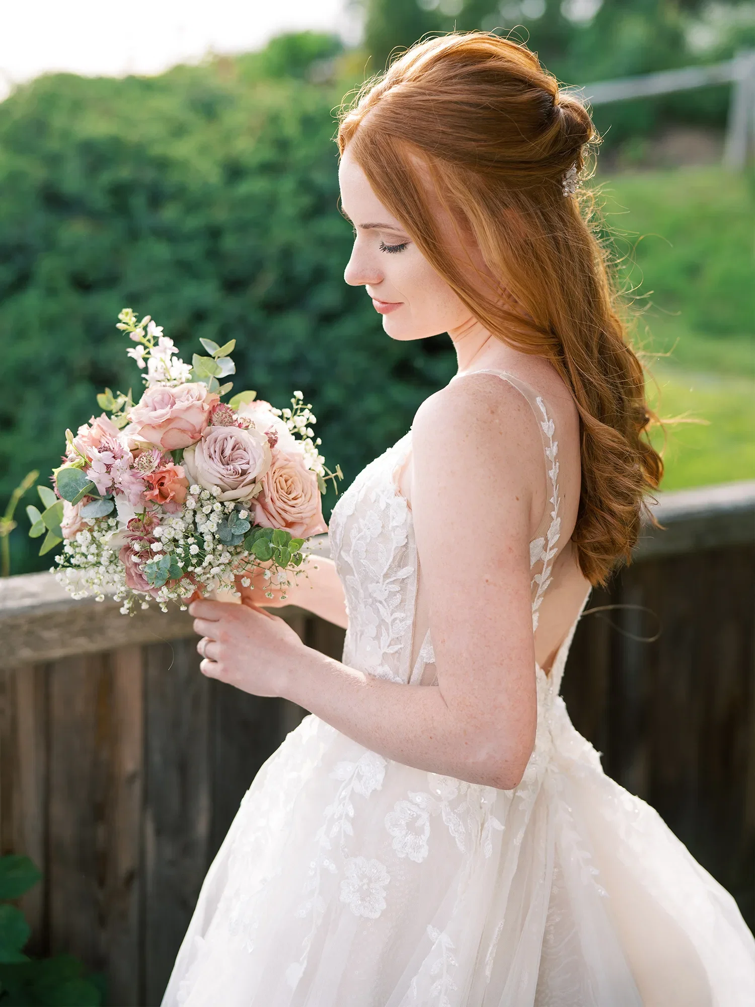 Elegante Braut mit roten Haaren, die in einem floralen Brautkleid und einem rosafarbenen Blumenstrauß in der Hand nach unten schaut, umgeben von Natur.